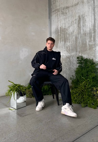 Chaqueta negra con cremallera y detalles blancos, camiseta negra, pantalones negros y zapatillas blancas. En un fondo de pared de concreto texturizado y vegetación.