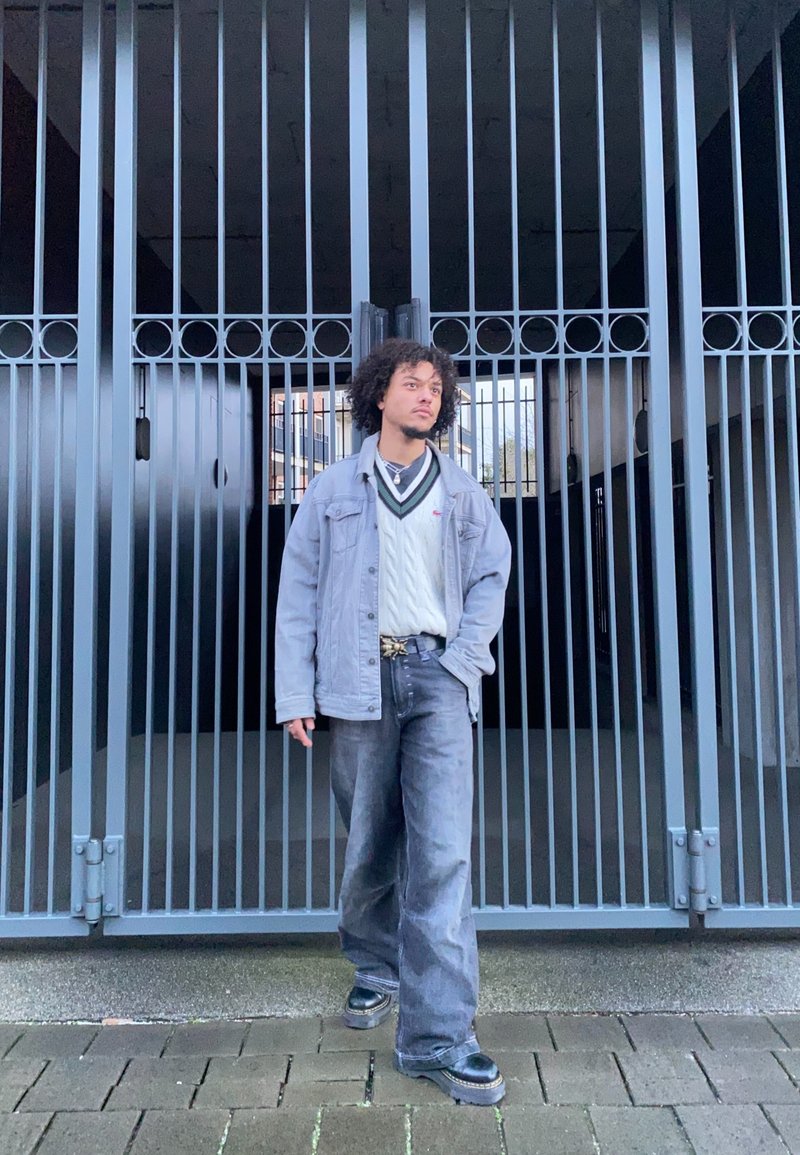 Szürke farmerdzseki egy fehér kötött pulóver felett, laza sötét farmernadrág és fekete platform cipő. A háttérben egy rácsos kapu látható.