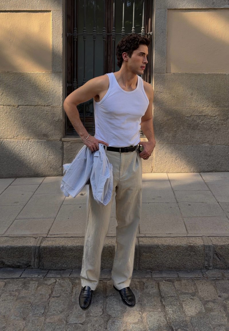 Jeune homme en débardeur blanc et pantalon beige se tient sur un trottoir en pierre en tenant une chemise rayée, avec un mur de pierre texturé derrière lui.