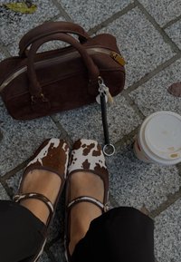 Balerinaskor med kobrorosttryck och bruna detaljer, en brun mockaväska med guldfärgade metallbeslag samt en vit kopp på en strukturerad stenyta.