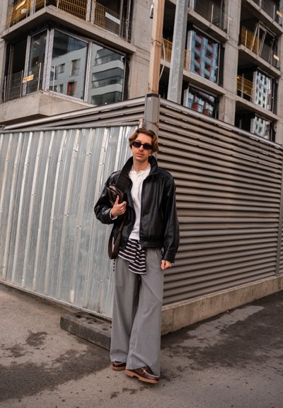 Joven con gafas de sol, chaqueta de cuero negra, pantalones holgados a rayas y zapatos marrones, de pie junto a barreras metálicas de construcción en un entorno urbano.