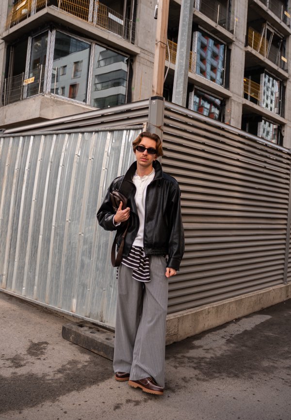 Νέος άνδρας με γυαλιά ηλίου, μαύρο δερμάτινο μπουφάν, φαρδιά ριγέ παντελόνια και καφέ παπούτσια στέκεται δίπλα σε μεταλλικά κατασκευαστικά φράγματα σε αστικό περιβάλλον.