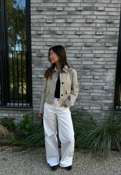 Chaqueta beige recortada con dos bolsillos frontales, combinada con pantalones de pierna ancha blancos. El fondo presenta una pared de ladrillo gris texturizada.