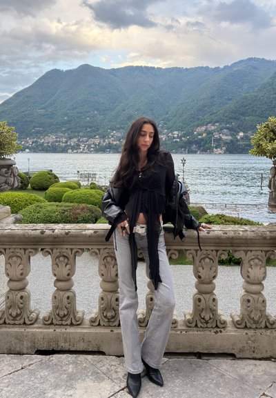 Mujer con cabello largo y oscuro, vestida con camiseta negra y vaqueros grises, se apoya en una barandilla de piedra con vistas a un lago y montañas verdes bajo un cielo nublado.