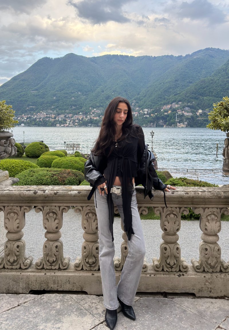 Femme aux longs cheveux foncés portant un haut noir et un jean gris, appuyée contre une rambarde en pierre avec vue sur un lac et des montagnes vertes sous un ciel nuageux.