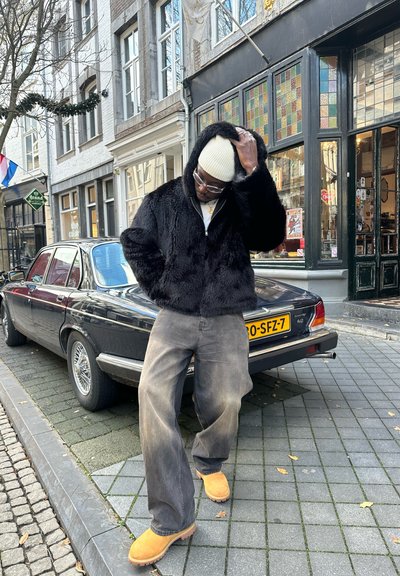 Chaqueta de piel sintética negra con capucha, jeans de pierna ancha grises, zuecos naranjas y un gorro crema. Coche vintage aparcado al fondo.