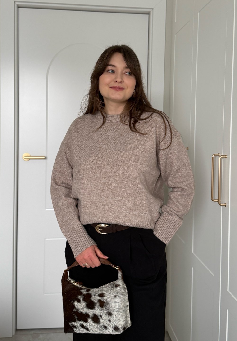 Femme portant un pull en maille beige et un pantalon noir, tenant un sac à main marron et blanc tacheté, debout à l'intérieur près de portes blanches et d'armoires.