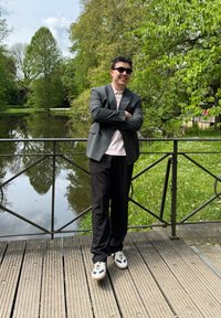 Un uomo in un blazer grigio sopra una camicia rosa chiaro, pantaloni neri e sneakers bianche si trova accanto a un parapetto vicino a uno stagno circondato da vegetazione.