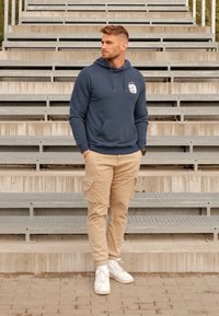 Granatowa bluza z kapturem z przednią kieszenią i sznurkami, w zestawieniu z beżowymi spodniami cargo z kieszeniami bocznymi oraz białymi trampkami.