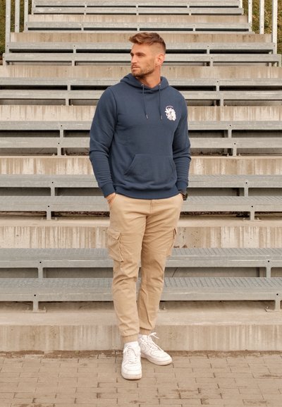 Sudadera con capucha azul marino con bolsillo frontal y cordones, combinada con pantalones cargo beige con bolsillos laterales y zapatillas blancas.