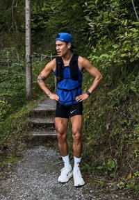 Idrettsutøver i blå tanktopp og svarte shorts, iført en svart løpevest. Hvite sko og sokker. Grønn skogbakgrunn med steintrapper.