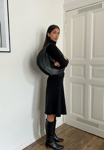 Bolso hobo de cuero negro colgado del hombro, combinado con un ajustado suéter negro de cuello alto y botas negras hasta la rodilla, contra un fondo neutro.