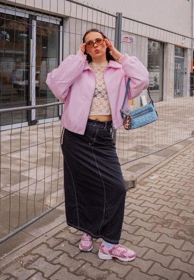 Mujer con chaqueta rosa, top corto de encaje, falda negra de rayas finas y zapatillas rosas posa con gafas de sol junto a una valla de obra.