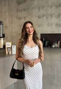 Biała sukienka w czarne kropki, dopasowany krój, sięgająca do połowy łydki. Czarna torebka z złotym okuciem, przezroczysty kubek z zimnym napojem, jasnoszare tło.