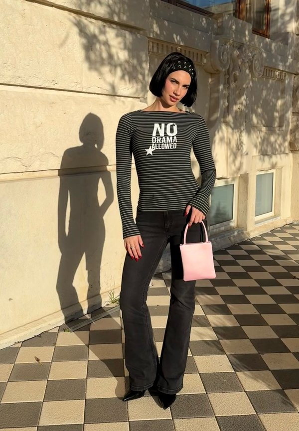 Femme aux cheveux courts noirs avec un bandeau, portant un haut rayé "No Drama Allowed", un jean noir, tenant un sac à main rose sur un passage pavé.