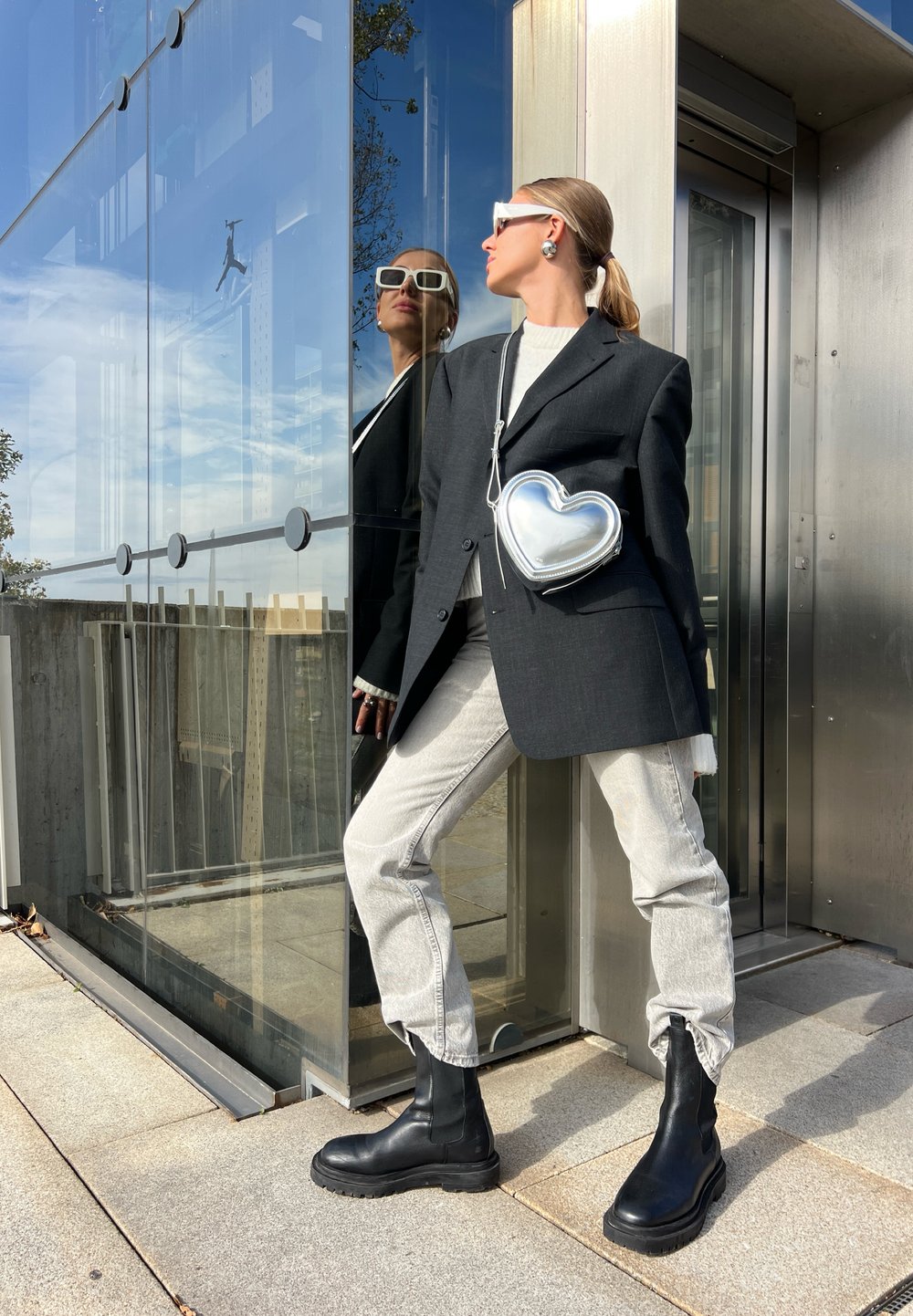 Svart kavaj, vit tröja, grå jeans och knähöga svarta stövlar. Silverfärgad hjärtformad väska. Reflekterande glasbyggnad i bakgrunden.