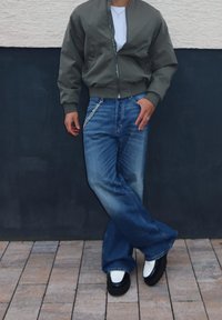 Casaco bomber verde-oliva, t-shirt branca, jeans flare azuis desbotados com detalhes em corrente e sapatos pretos com detalhes brancos.