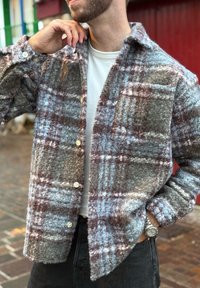 Flaneļa krekls pelēkā, zilā un brūnā pleda rakstā; raksturo teksturēts audums, pogu aizdare un apkakle; modelis to valkā virs baltas blūzes.