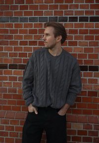 Mies, jolla on kädetTaskuissa, päällään harmaa palmikkoneule ja mustat housut, seisoo punaisen tiiliseinän edessä katsomassa sivulle.