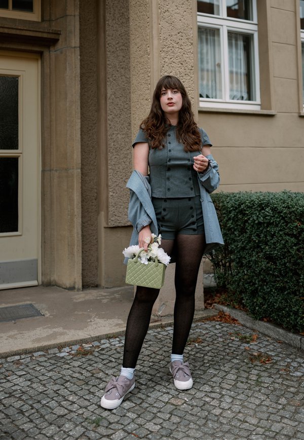 Mujer con pelo largo y castaño, de pie al aire libre, lleva una camiseta gris abotonada y pantalones cortos, medias negras, chaqueta gris, zapatillas deportivas, sosteniendo una bolsa verde con flores blancas.