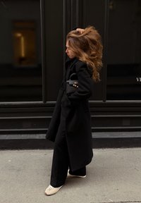 Kvinde i sort frakke bærer en sort håndtaske, står sidelæns på fortovet, løfter langt bølget hår med den ene hånd, iført beige sko.