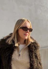 Rjavi bikačasti sončni očala, mehak bež pulover z zadrgo in temen puhast zunanji kos oblačila. Naravna svetloba poudarja teksture.