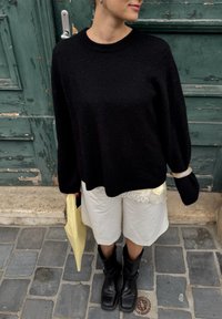 Svart stickad tröja med långa ärmar, ihop med vita shorts. Svarta ankelstövlar och en ljusgul väska syns, mot en grön bakgrund.