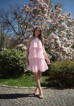 Vestido de un suave tono rosa pálido, con capas en forma de volantes y un escote halter. Combinado con un pequeño bolso blanco.