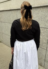 Mujer con cabello rubio largo sujeto con un gran lazo de terciopelo negro, vistiendo una blusa negra y una falda blanca, de pie cerca de una pared de piedra gris.