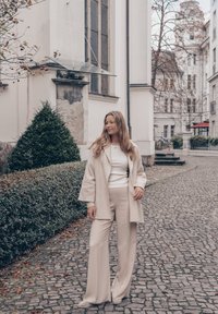 Manteau en laine beige porté sur un pull blanc, associé à un pantalon large beige. Texture lisse et coupe ample, sur fond de chemin pavé.