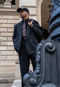 Suit grigio scuro doppiopetto con motivo verticale, abbinato a una camicia nera, cravatta a righe, occhiali da sole e un berretto nero. Tiene una borsa.
