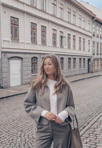 Grå jakke i ullblanding med vide ermer, over en hvit kabelstrikket genser. Grå bukser og en myk grå håndveske. Bakgrunn med brosteinsgate.