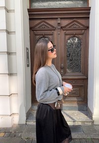 Szary sweter z długimi rękawami, czarna spódnica oraz akcesoria, w tym biały bransoletka i mała brązowa torebka na pasek. Trzyma kubek.