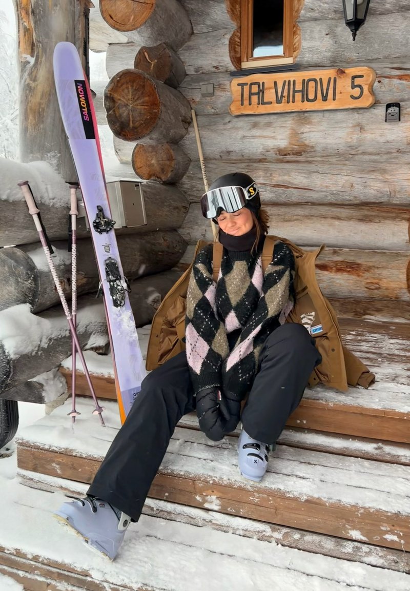 Personne en tenue de ski assise sur les marches enneigées d'une cabine à côté de skis et bâtons, panneau en bois portant l'inscription "TALVIHOVI 5" sur le mur en rondins derrière.