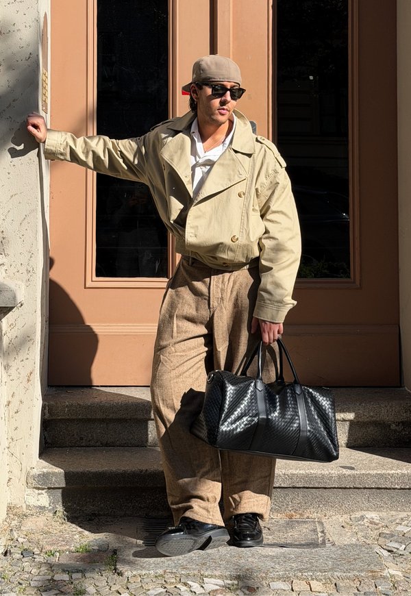 Hombre con chaqueta beige, pantalones marrones, gorra hacia atrás y gafas de sol, sosteniendo una bolsa negra, apoyado en un marco de puerta al aire libre.