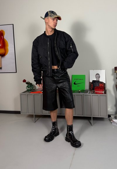 Chaqueta bomber negra, top negro corto, pantalones cortos de cuero y botas negras hasta la espinilla. Presenta una gorra desgastada azul y roja.