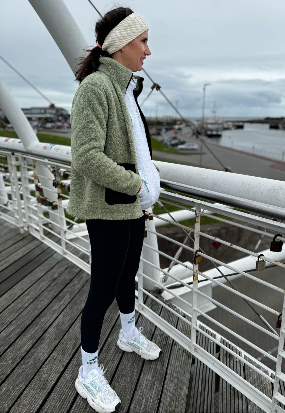 Veste en polaire vert clair sur un sweat à capuche gris, leggings noirs et chaussures de sport blanches. Debout sur un pont avec une rivière et des voitures stationnées en arrière-plan.
