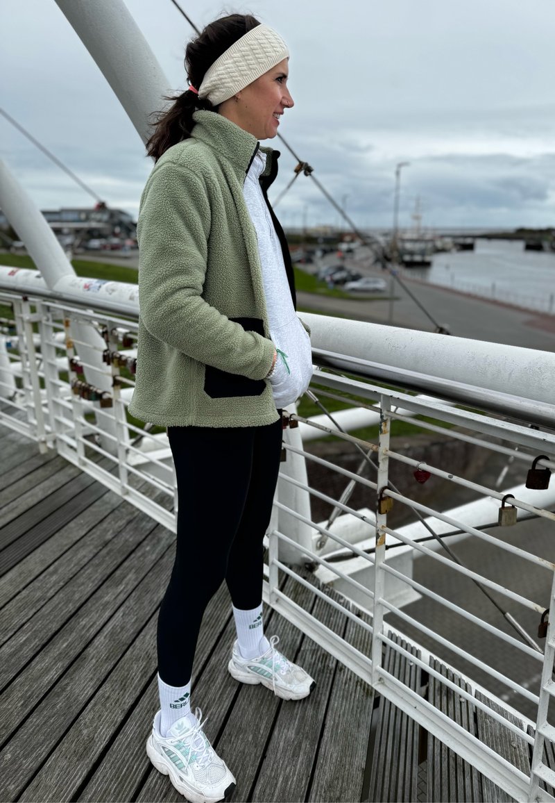 Veste en polaire vert clair sur un sweat à capuche gris, leggings noirs et chaussures de sport blanches. Debout sur un pont avec une rivière et des voitures stationnées en arrière-plan.
