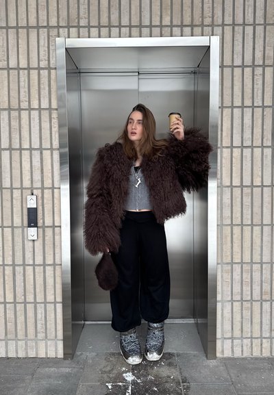 Joven con chaqueta marrón esponjosa sosteniendo una taza de café, de pie dentro de un ascensor de acero inoxidable con paredes de baldosas grises.