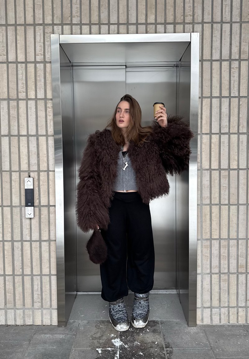 Junge Person in einer flauschigen braunen Jacke, die eine Kaffeetasse hält, steht in einem Aufzug aus Edelstahl mit grauen Fliesenwänden.