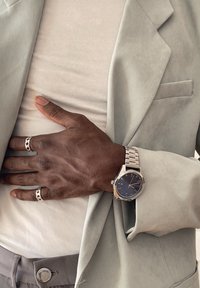 Tmavá koža na ruke s striebornými prsteňmi a striebornými hodinkami spočíva na svetlo béžovom saku a bielej košeli nad sivými nohavicami.