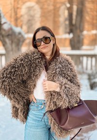 Manteau en fourrure marron pelucheux, haut blanc côtelé, jean taille haute bleu clair, sac à main en cuir bordeaux foncé, lunettes de soleil noires surdimensionnées. Fond enneigé.