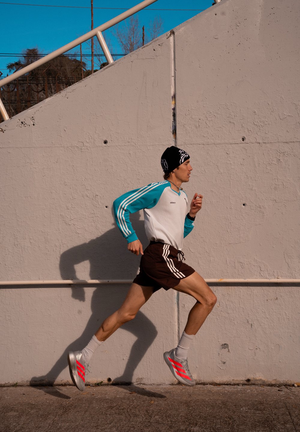 Coureur portant une chemise à manches longues bleu sarcelle et blanche, un bonnet noir, un short noir et des chaussures argentées avec des rayures rouges courant à côté d'un mur en béton gris.