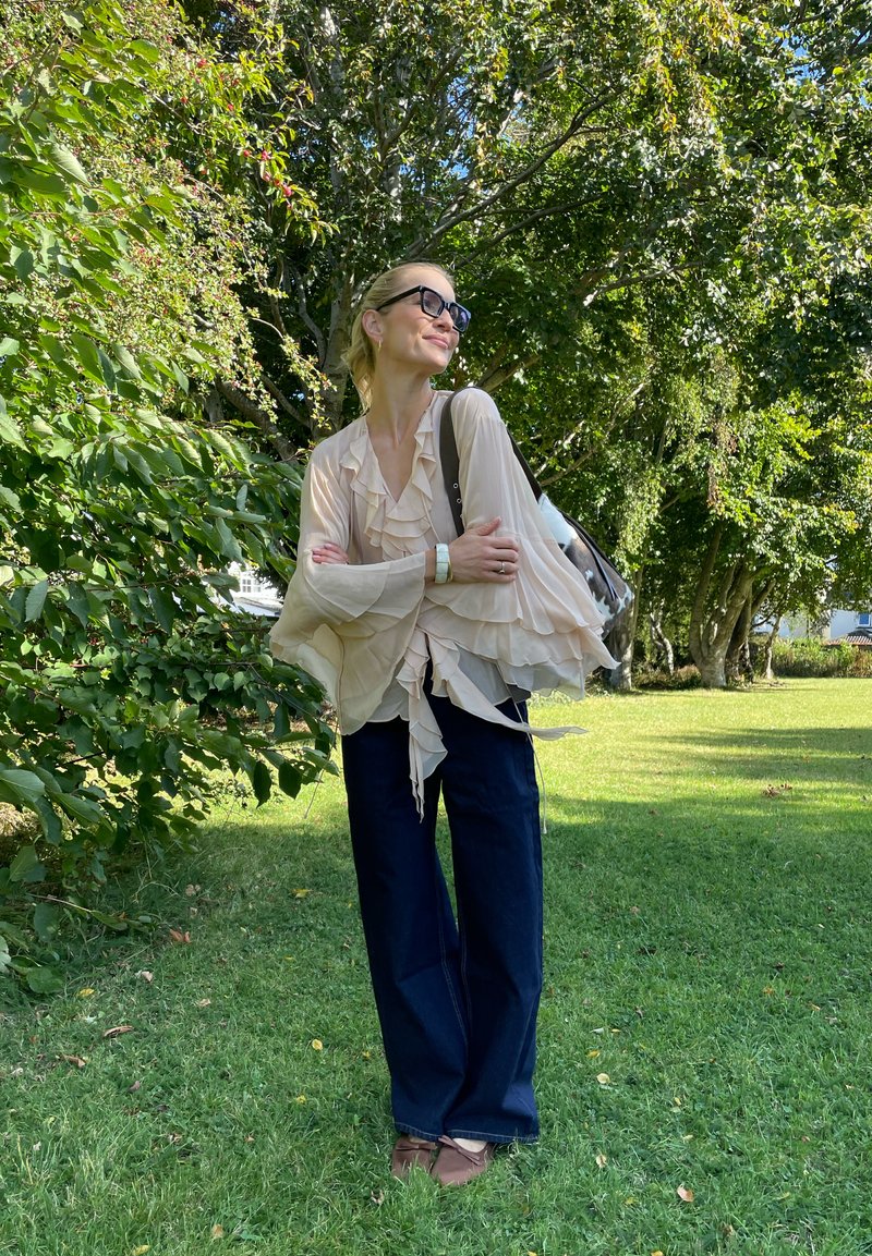 Bluse in hellbeige mit Rüschen und langen Ärmeln, kombiniert mit dunkelblauen weiten Jeans und braunen Schuhen. Das Model steht in einer grünen Außenumgebung.