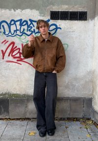 Hnědá semišová bunda na zip s elastickými manžetami, kombinovaná s volnými černými džínami a černými botami. Na pozadí je graffiti na zdi.