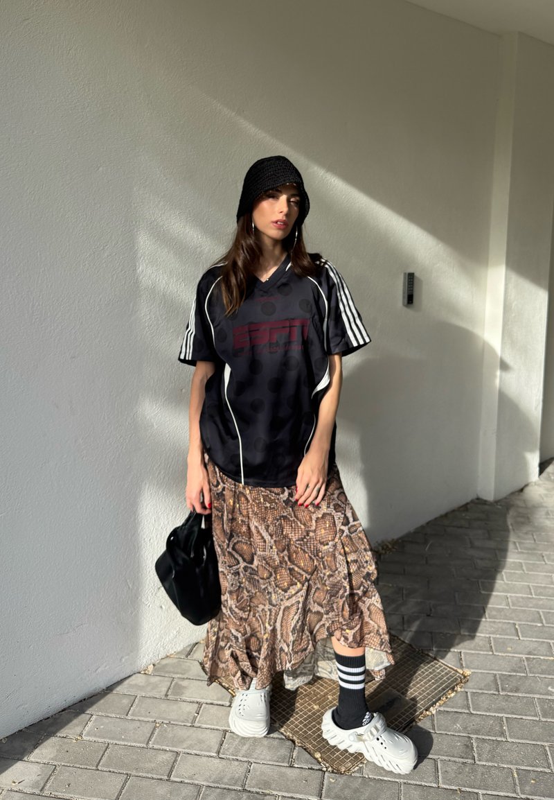 Femme portant un chapeau bob noir, un maillot de sport noir, une jupe à imprimé serpent, des chaussettes rayées, des chaussures blanches épaisses, et tenant un sac noir, debout sur le trottoir.