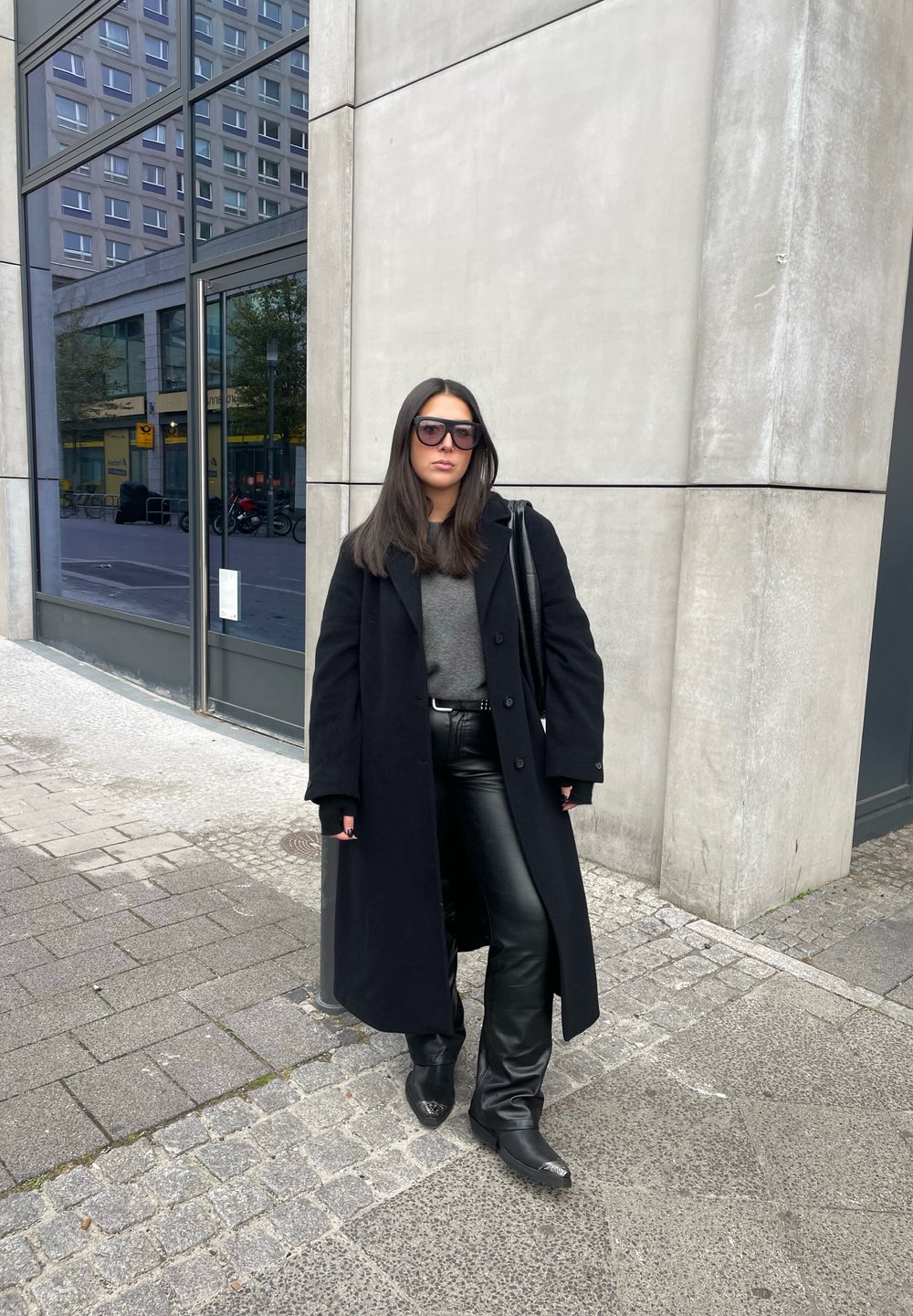 Μαύρο μάλλινο παλτό πάνω από γκρι πουλόβερ, σε συνδυασμό με μαύρα δερμάτινα παντελόνια και τακούνια. Υπερμεγέθη γυαλιά ηλίου, δημόσιος αστικός χώρος.