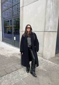 Μαύρο μάλλινο παλτό πάνω από γκρι πουλόβερ, σε συνδυασμό με μαύρα δερμάτινα παντελόνια και τακούνια. Υπερμεγέθη γυαλιά ηλίου, δημόσιος αστικός χώρος.