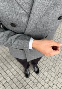 Szary płaszcz w jodełkę z podwójnym zapięciem oraz czarnymi guzikami, w połączeniu z białą koszulą i czarnymi butami; stojący na brukowanej powierzchni.