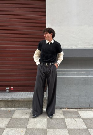 Suéter negro con textura sobre una camisa blanca de cuello, combinado con pantalones grises de cintura alta y zapatos negros; posando frente a un fondo marrón.